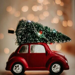 A red toy car carries a Christmas tree, set against a warm bokeh background, celebrating the festive season.