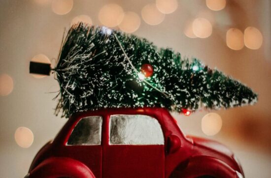 A red toy car carries a Christmas tree, set against a warm bokeh background, celebrating the festive season.