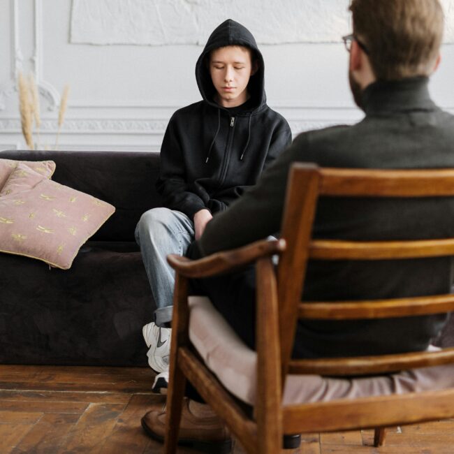 A teenager in hooded sweatshirt talks with a therapist in a modern, minimalistic studio.