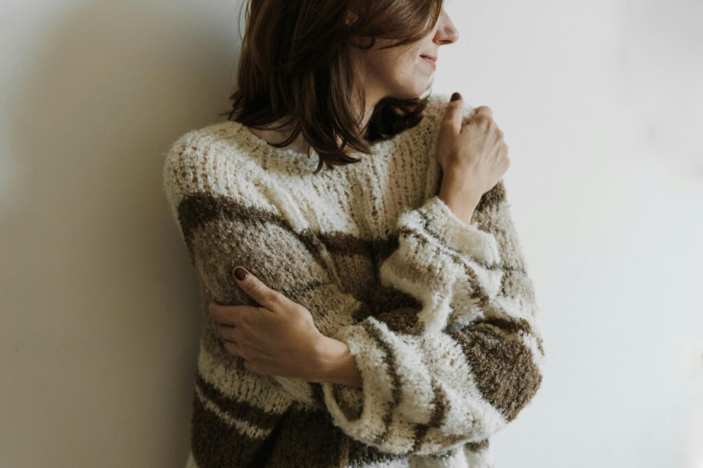 Woman in a knit sweater hugging herself with a serene expression, indoors.