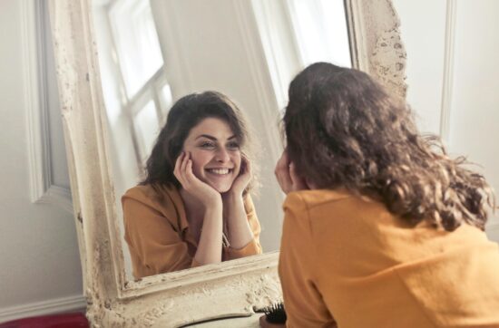 A cheerful woman smiles at her reflection in a vintage-style mirror, exuding positivity and warmth.